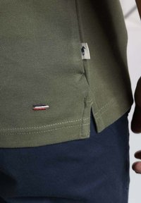 Polo shirt vert olive avec un tissu texturé, des fentes sur les côtés et un petit logo brodé en rouge, blanc et bleu près de l'ourlet.