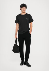 Fekete pamut póló, kis grafikával a mellrészen, fekete melegítő nadrággal és fényes fekete lakkcipővel párosítva. Oldaltáska a kézben.