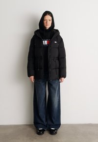 Musta puhvijakku, jossa on huppu, vetoketju edessä ja kiristettävä vyötärö, mustan hupparin päällä, jossa on monivärinen logo, yhdistettynä leveälahkeisiin farkkuihin.