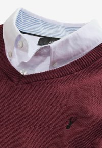 Pull bordeaux en tissu texturé, design à col en V et logo noir. Col de chemise blanche avec des accents rayés bleus en dessous.