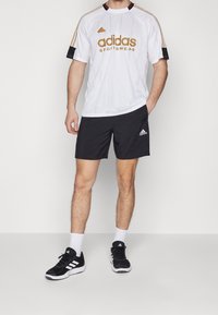 Vitt kortärmat jersey med guld "adidas SPORTSWEAR"-logotyp, svarta detaljer, tillsammans med svarta shorts och svarta sneakers med vita ränder.