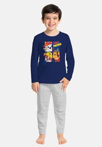 Ragazzo sorridente che indossa una maglietta a maniche lunghe navy blu di Paw Patrol e pantaloni grigio chiaro, in piedi scalzo su uno sfondo neutro.