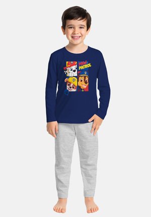 Smiling boy wearing a navy blue long-sleeve Paw Patrol shirt and light gray pants, standing barefoot against a plain background.