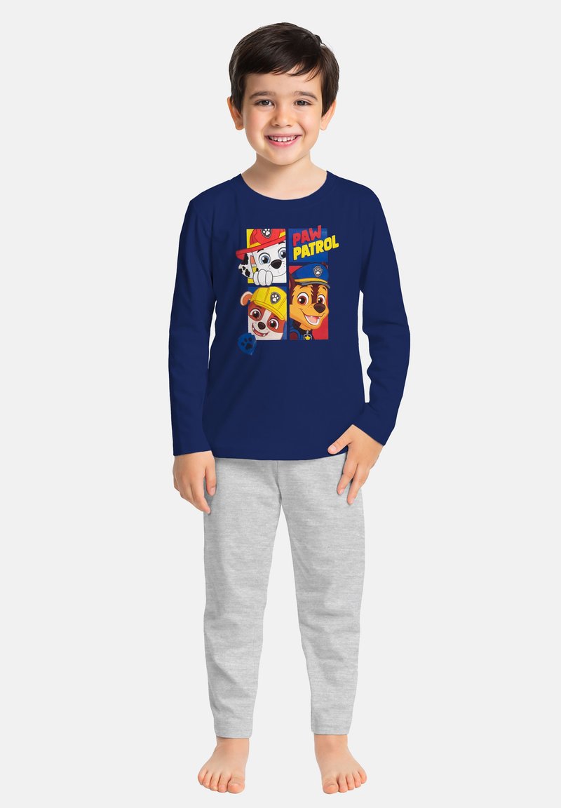 Ragazzo sorridente che indossa una maglietta a maniche lunghe navy blu di Paw Patrol e pantaloni grigio chiaro, in piedi scalzo su uno sfondo neutro.