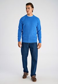 Niebieski sweter w sploty z okrągłym dekoltem i ściągaczami na rękawach, w zestawieniu z ciemnoniebieskimi dżinsami i brązowymi sznurowanymi butami.