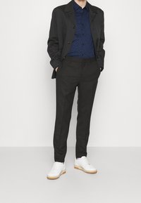 Svart kavaj över en blåmönstrad skjorta, kombinerad med svarta byxor och vita sneakers. Utstrålar en modern, skräddarsydd stil.