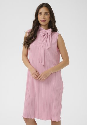 Kobieta o długich ciemnych włosach, ubrana w różową plisowaną sukienkę bez rękawów z kokardą przy szyi, stojąca na jednolitym jasnym tle.