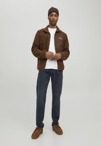 Chaqueta de forro polar marrón con cremallera, bolsillos laterales y un ligero cuello, llevada sobre una camiseta blanca, combinada con unos jeans azul oscuro y zapatos marrones.