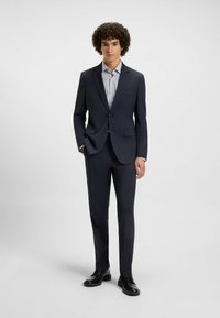 Marineblauw kostuum met een getailleerde pasvorm, voorzien van een blazer met twee knopen, vlakke broek en een lichtblauw gestreept overhemd. Glanzend zwarte schoenen.
