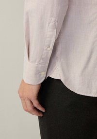 Camisa a rayas en rosa claro y blanco, con puños abotonados y tejido suave. El dobladillo es ligeramente curvado, descansando sobre pantalones oscuros.