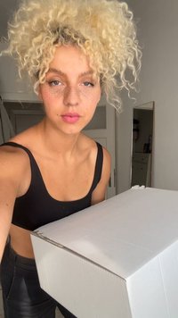 Weißer Karton, der vor einer Person mit lockigem, blonden Haar gehalten wird. Die Box hat eine glatte Oberfläche und eine flache Oberseite, die sich gut zum Versand eignet.