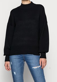Schwarzer Strickpullover mit hohem Kragen, überschnittenen Schultern und gerippten Bündchen. Kombiniert mit ausgebeulten blauen Skinny-Jeans.