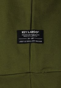 Tessuto verde oliva con una maglia testurizzata. Etichetta nera con testo bianco che indica "KEY LARGO", logo e dettagli del prodotto. Orlo a coste.