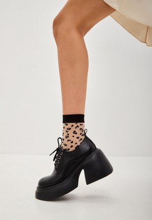 Zapatos de plataforma negros con punta redonda y detalle de cordones, combinados con calcetines hasta el tobillo de lunares que tienen un diseño de estampado de leopardo.