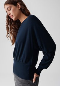 Navyblaues Langarmshirt aus weichem Stoff, mit lockerer Passform und runder Taille. Merkmale sind ein V-Ausschnitt und weite Ärmel, kombiniert mit grauen Jeans.