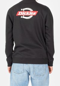 Sweatshirt noir avec poignets et ourlet côtelés, arborant un grand logo rouge "Dickies" dans le dos. Porté avec un jean en denim bleu clair.