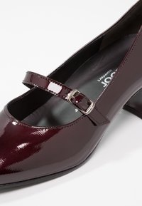 Chaussure Mary Jane en cuir verni bordeaux avec bout rond, bride à boucle métallique réglable et un talon de hauteur intermédiaire, dotée d'une finition lisse.