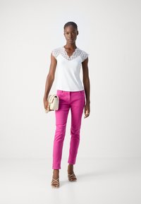 Haut blanc avec des détails en dentelle aux épaules, associé à un pantalon rose ajusté. Le mannequin tient un sac à main beige et porte des sandales à brides.
