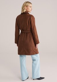 Palton maro din piele întoarsă, cu curea, guler și mâneci lungi, asortat cu blugi largi, culoare bleu deschis, și cizme negre cu vârful ascuțit.