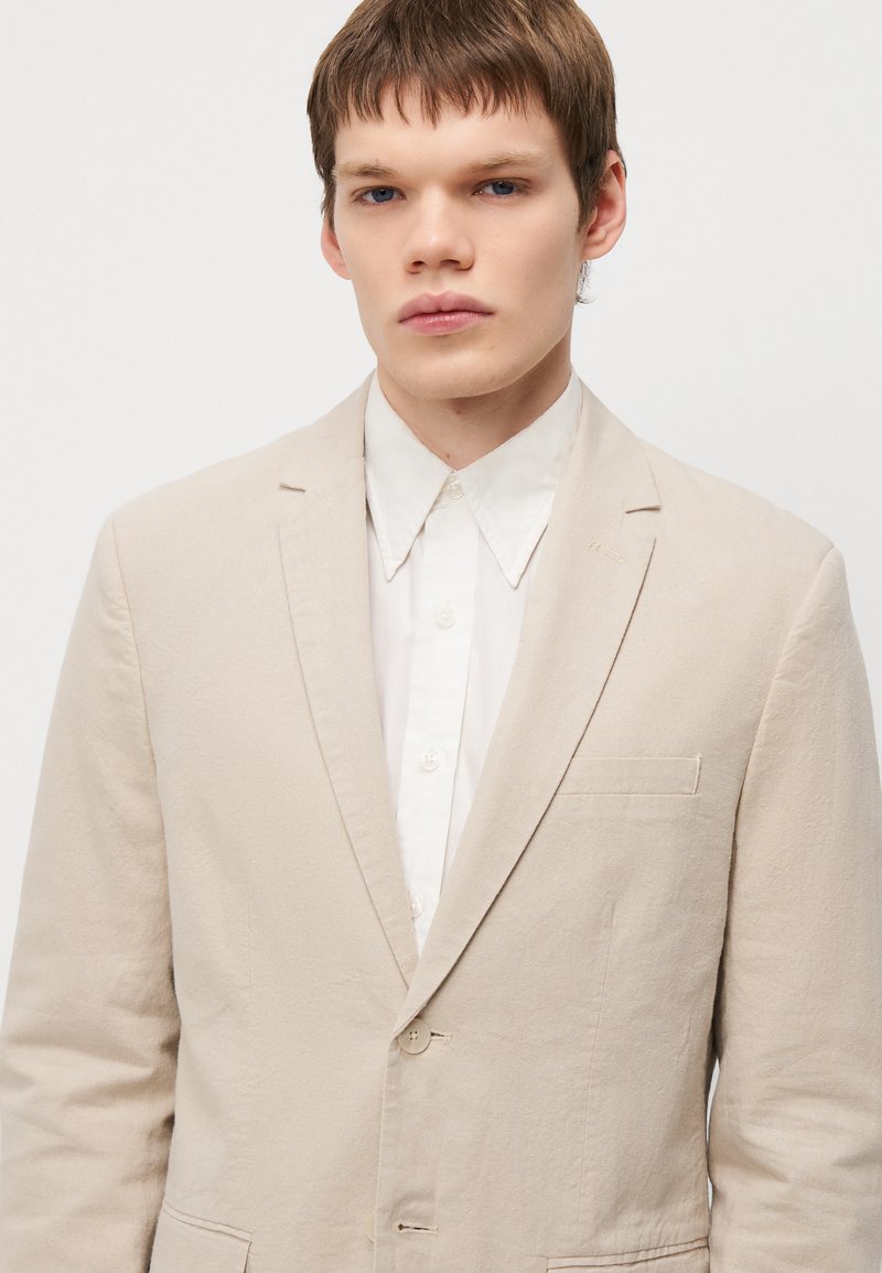 Blazer beige clair en tissu texturé, avec deux boutons, une petite poche poitrine et des revers crantés, superposé sur une chemise blanche à boutons.
