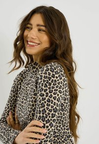 Haut à manches longues imprimé léopard en beige et noir avec des accents violets ; tissu doux, coupe ajustée, et col montant. Le mannequin pose avec les bras croisés.
