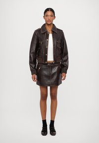 Giacca cropped in pelle marrone con tasche frontali e cuciture a contrasto, abbinata a una gonna in pelle coordinata con cerniere.