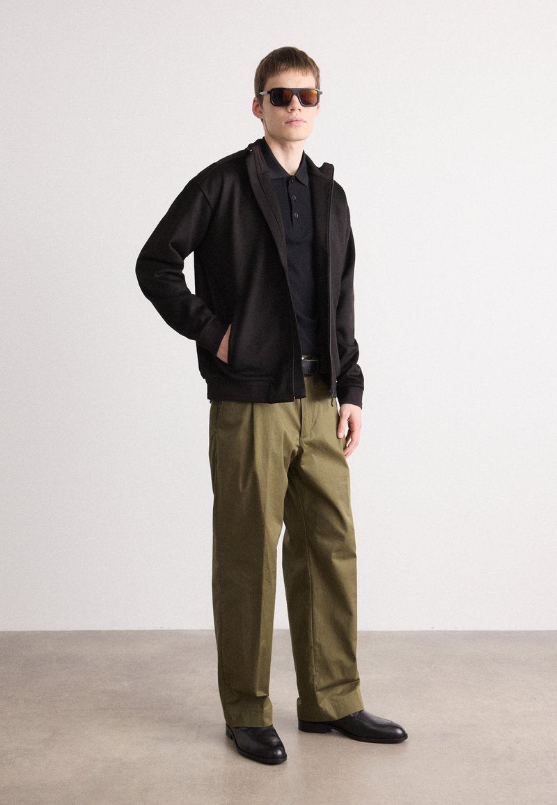 Schwarze Zip-Up-Jacke, schwarzes Poloshirt, olivgrüne Hose und schwarze Schuhe. Quadratische Sonnenbrille mit braunen Gläsern. Schlicher Hintergrund.
