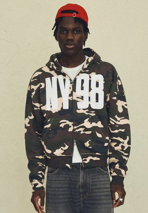 Junger Mann mit roter Kappe und Camouflage-Hoodie mit "NY 98" großen weißen Buchstaben, der vor einer schlichten hellen Wand steht.