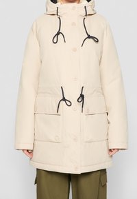 Parka beige isolée avec capuche, cordon de serrage et quatre boutons. Comprend deux poches avant et des accents de cordon de serrage noirs.