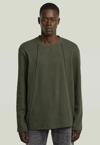 Homme portant un pull long à manches longues de couleur vert olive texturé avec des lignes de couture verticales visibles, associé à un jean foncé décoloré, se tenant devant un fond uni.