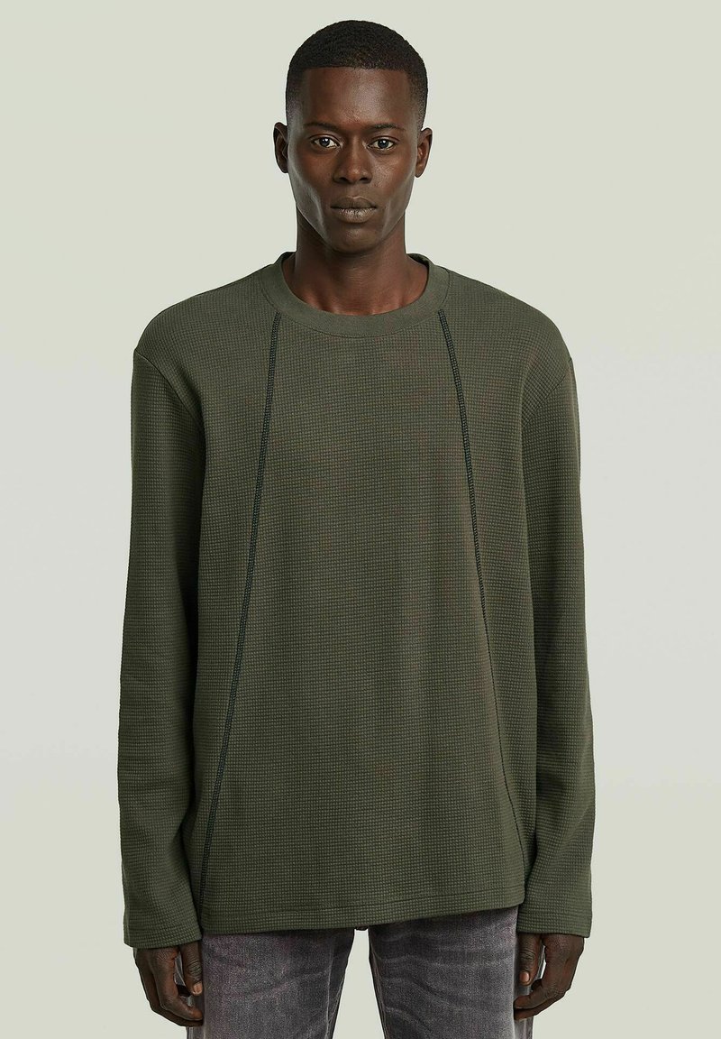 Homme portant un pull long à manches longues de couleur vert olive texturé avec des lignes de couture verticales visibles, associé à un jean foncé décoloré, se tenant devant un fond uni.
