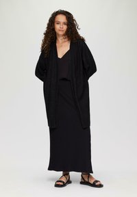 Langes schwarzes Kleid mit V-Ausschnitt, kombiniert mit einer locker sitzenden schwarzen Strickjacke. Strukturiertes Strickmaterial und schwarze Sandalen runden das Outfit ab.