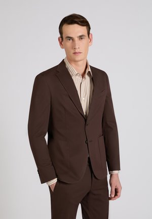 Man die een donkerbruin pak en een beige gestreept overhemd draagt, staand tegen een effen lichtgrijze achtergrond.