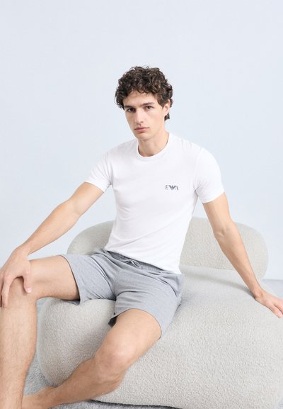 Balta kokvilnas T-krekls ar mazu logotipu uz krūtīm, kopā ar pelēkiem šortiem. Modelis sēž uz fakturētas bēšas krēsla gaišā vidē.