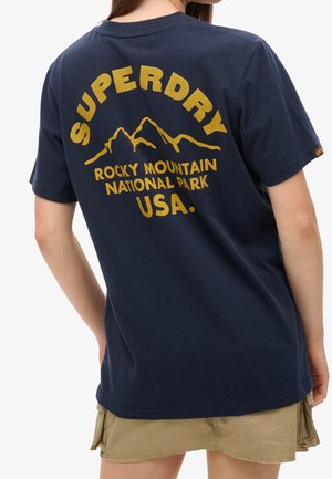 Osoba nosząca granatową koszulkę Superdry z żółtym napisem Rocky Mountain National Park USA i grafiką gór na plecach.