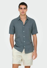Camisa de botones de manga corta en un tono verde azulado oscuro, hecha de una tela ligera, combinada con pantalones cortos de un beige claro, que muestran un ajuste relajado y un cuello.