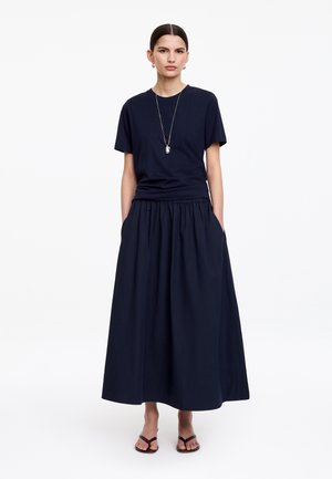 Žena stojí oblečená v tmavomodrom krátkokopevnom tričku, dlhú nariasenú sukňu s vreckami, čierne sandále a dlhý príveskový náhrdelník na bielom pozadí.
