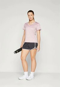 O tânără îmbrăcată cu o bluză roz cu guler în V, pantaloni scurți negri de sport, șosete albe și adidași albi, ținând o sticlă de apă neagră, stând în fața unui fundal simplu.