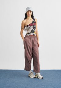 Casquette grise, débardeur graphique asymétrique avec texte et image, pantalon ample rose poussiéreux et baskets beiges avec accents foncés.