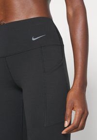 Gros plan d'une personne portant des leggings noirs Nike avec une poche latérale et une partie médiane et une main visibles sur un fond clair uni.