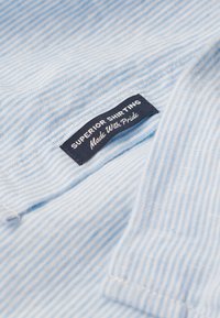 Lichtblauwe stof met horizontale witte strepen, voorzien van een zwart label met de tekst "SUPERIOR SHIRTING Made With Pride."