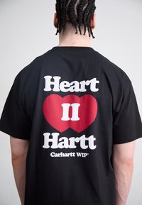 Zwarte katoenen T-shirt met een grote rode hartafbeelding en de tekst "Heart II Hartt" in vette witte letters. Inclusief Carhartt WIP® branding.
