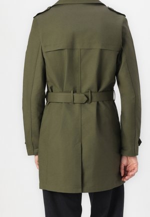Vue de dos d'une personne portant un trench-coat vert olive ceinturé à manches longues sur un pantalon noir.