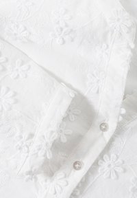 Valkoinen broderi-paita, jossa on kukkakuvioita, teksturoitua kangasta ja pyöreitä nappeja, joissa on kukkaaksentteja läpällä.