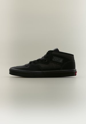 Zapatilla negra de caña media con cordones delanteros, costuras visibles y etiqueta "Half Cab" en el lateral, mostrada sobre un fondo claro y liso.