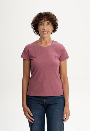Femme aux cheveux bruns courts et bouclés portant un t-shirt rose et un jean bleu, debout et souriante devant un fond clair uni.