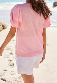 Suéter rosa claro con mangas abullonadas, dobladillo acanalado y detalle texturizado en el cuello, combinado con pantalones cortos de lino blancos en una playa de arena.