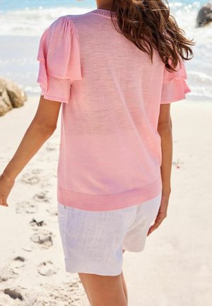 Jasnoróżowy sweter z bufkami, prążkowanym dołem i teksturowym detalem przy dekolcie, w połączeniu z białymi lnianymi szortami na piaszczystej plaży.