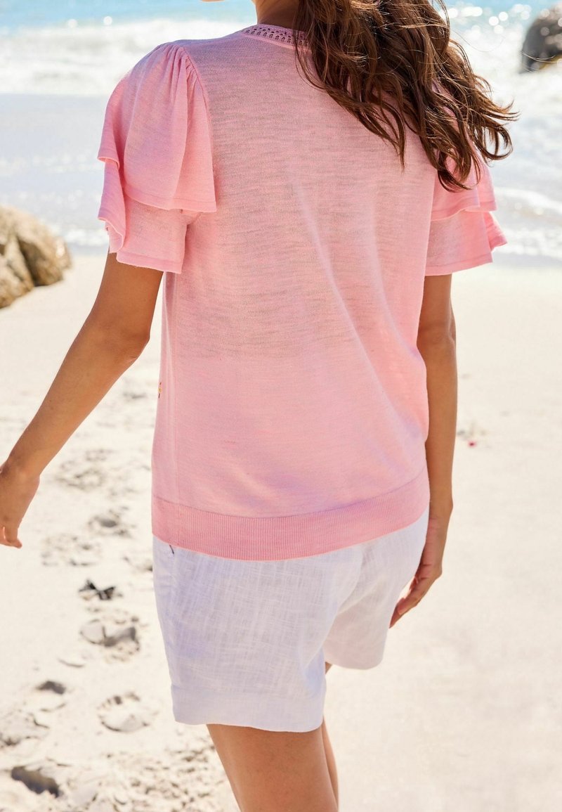 Suéter rosa claro con mangas abullonadas, dobladillo acanalado y detalle texturizado en el cuello, combinado con pantalones cortos de lino blancos en una playa de arena.