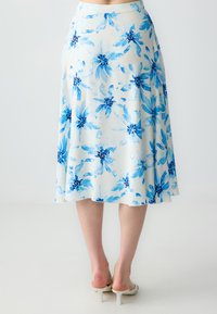 Donna che indossa una gonna midi bianca con stampa floreale blu e sandali con tacco bianchi, vista dalla vita in giù su uno sfondo neutro.